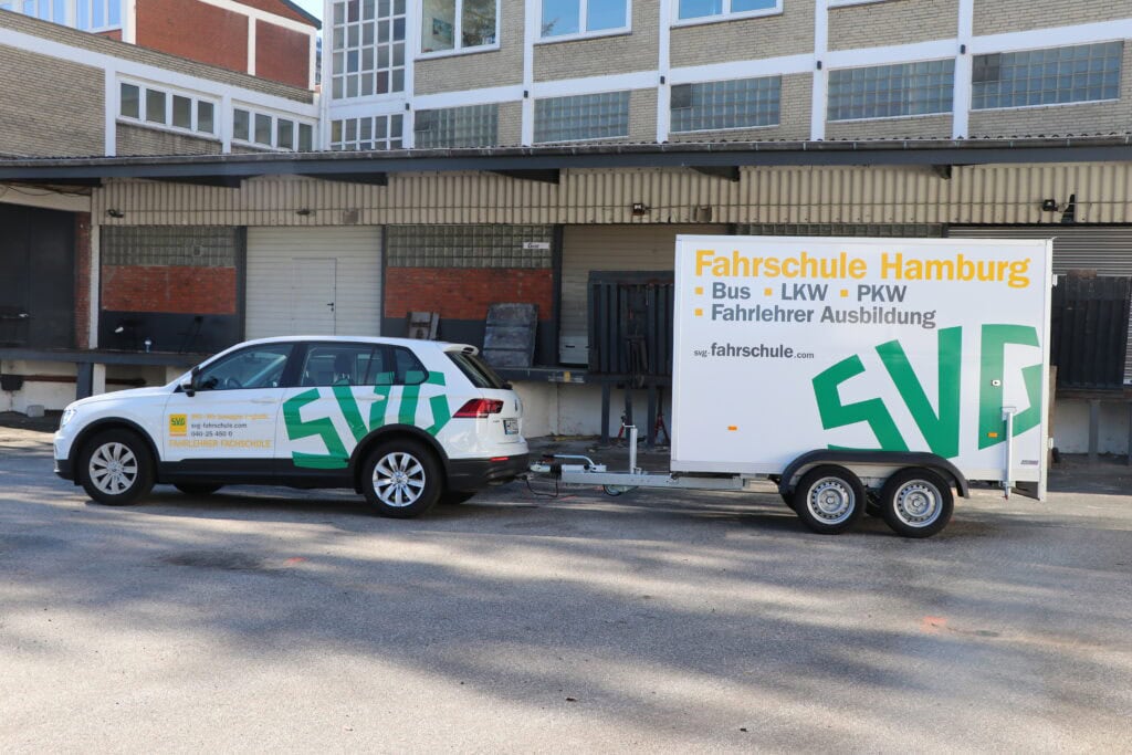 Auto mit Anhänger, Fahrschule Hamburg-Werbung, weiß, steht vor einem Industriegebäude.