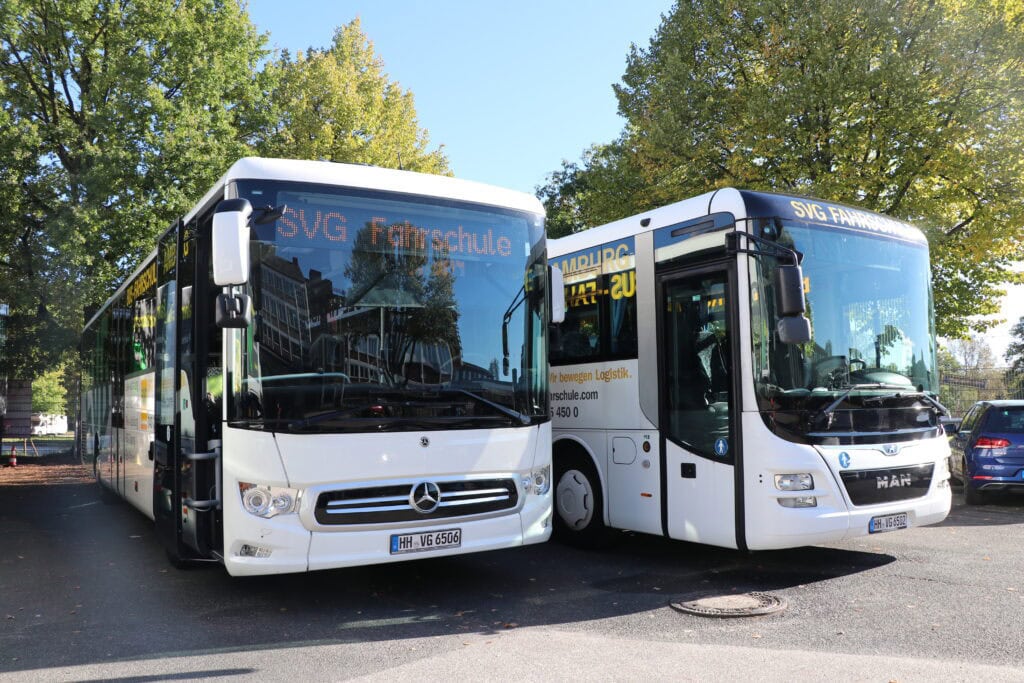 Zwei weiße Schulbusse, Mercedes und MAN, bei Tageslicht, geparkte Bäume im Hintergrund.